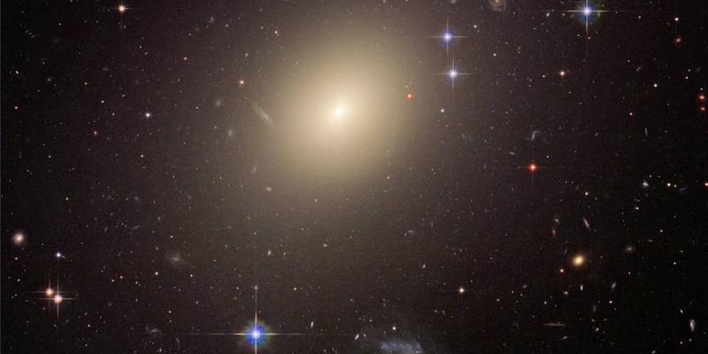 Una grande galassia ovale gialla si trova in un campo pieno di molte galassie più piccole. Questa grande galassia è più luminosa nel suo nucleo