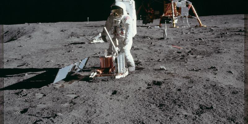 An astronaut in a white spacesuit stands on the grey lunar surface with a piece of equipment in-front of a lunar lander