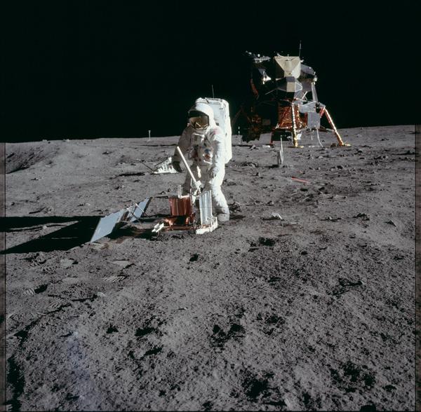 Un astronauta in tuta bianca dispone dell'attrezzatura sul grigio terreno lunare a pochi passi dal modulo di discesa