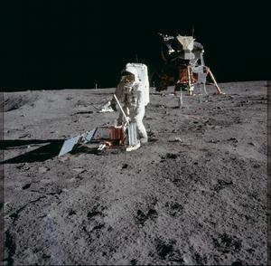 An astronaut in a white spacesuit stands on the grey lunar surface with a piece of equipment in-front of a lunar lander