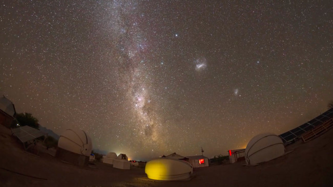 Over a cluster of small telescope domes the Milky Way juts upward from the horizon. Two fuzzy blobs are on the right