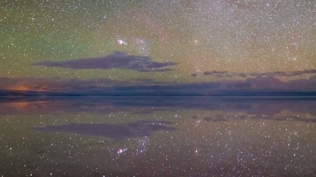 In a field of countless stars dotted by clouds and reflected in water, the three stars of Orion’s belt poke above the horizon