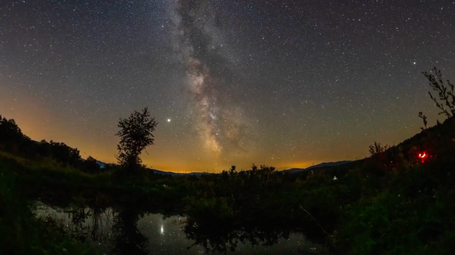 La Via Lattea si staglia all'orizzonte su un paesaggio con alberi, acqua e il lontano bagliore delle luci della città.