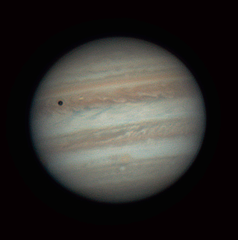 Jupiter with coloured horizontal bands of clouds. The shadow of the moon Io is seen as a dark circle in the top left