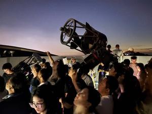 Several people looking up at the sky, two medium-sized telescopes are amongst the crowd