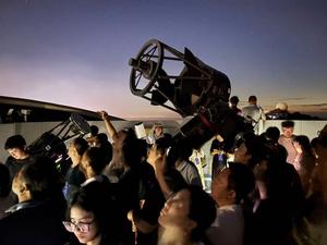 Several people looking up at the sky, two medium-sized telescopes are amongst the crowd