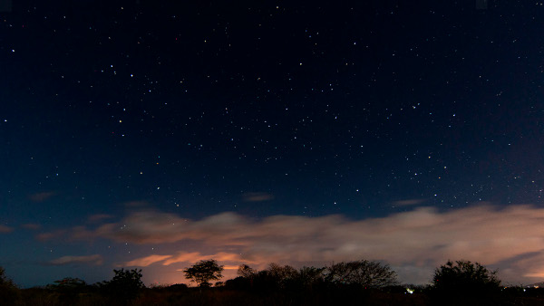 A starry sky over a cloudy landscape. On the left the bright red star Antares sits at the top of a hook-shaped star pattern