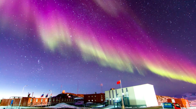 A Y-shaped band of light with light and dark patches, in the colours green and pink glows in the sky over some buildings