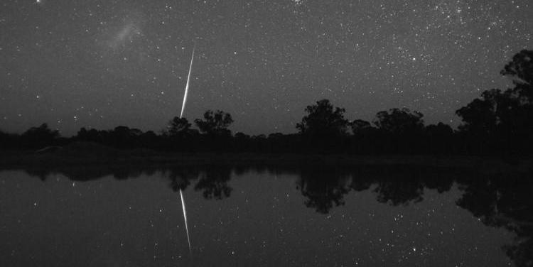 The bright streak of a meteor is reflected in a body of water. The Large Magellenic Cloud is in the top left as a fuzzy line