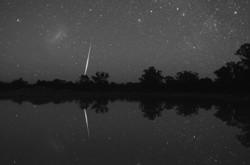 The bright streak of a meteor is reflected in a body of water. The Large Magellenic Cloud is in the top left as a fuzzy line