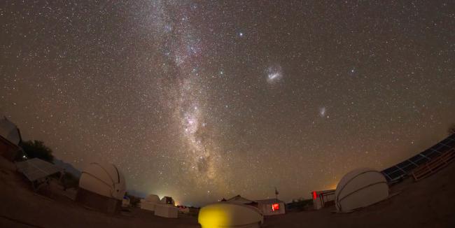 Over a cluster of small telescope domes the Milky Way juts upward from the horizon. Two fuzzy blobs are on the right