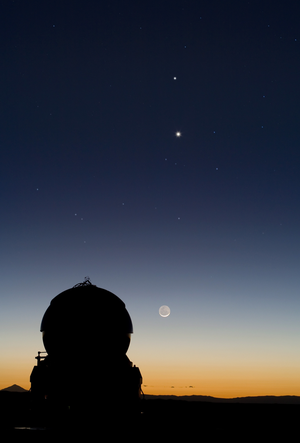 The crescent Moon with Venus & Mercury above it in a line in a bright twilight sky above a silhouetted telescope dome