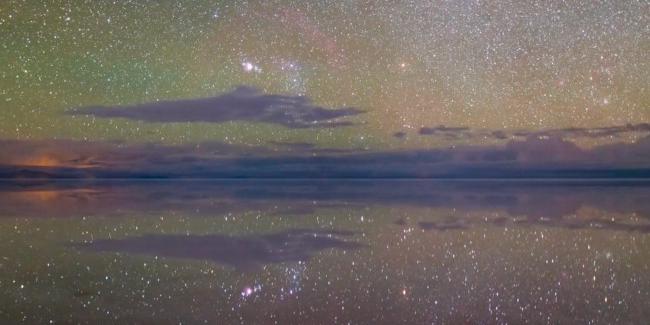 In a field of countless stars dotted by clouds and reflected in water, the three stars of Orion’s belt poke above the horizon
