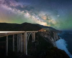 The Milky Way looms over a bridge on a steep coastline. To the upper right is the bright, deep red star Antares