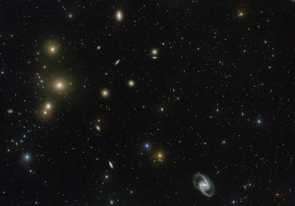 Raggruppamento sparso di galassie con molte galassie ellittiche giallastre e una galassia a spirale in primo piano.