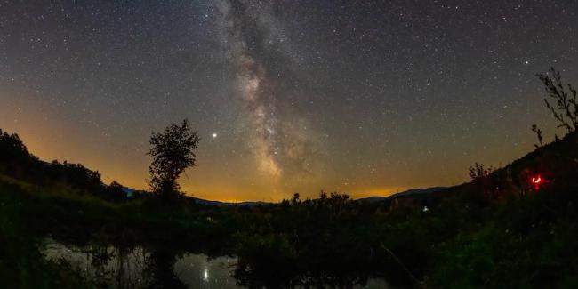 The Milky Way rises from the horizon over a landscape with trees, water and the distant glow of city lights