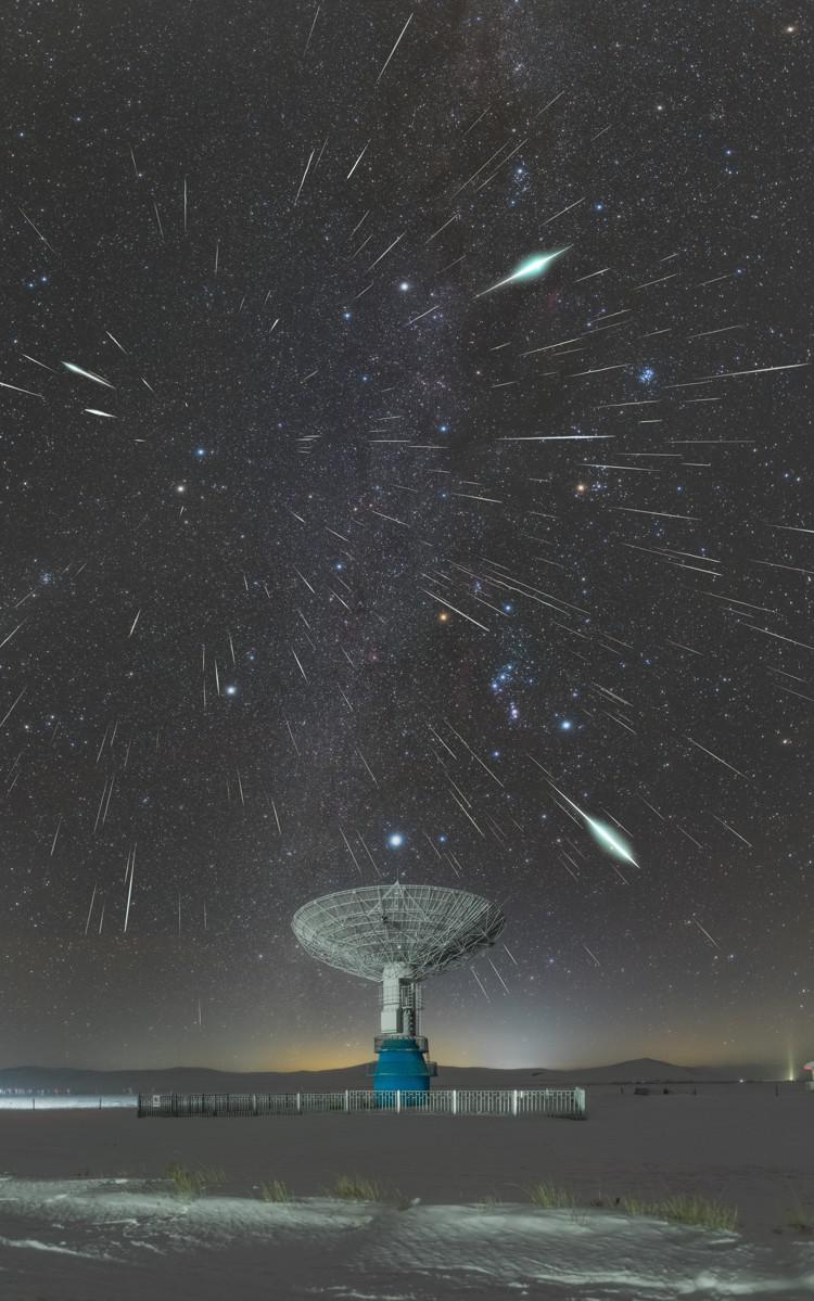 Bright streaks created by meteors radiate from a point in the sky above the dish of a radio telescope.