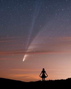 La sagome di una donna sembra salutare una cometa che appare dietro strati di nuvole leggere