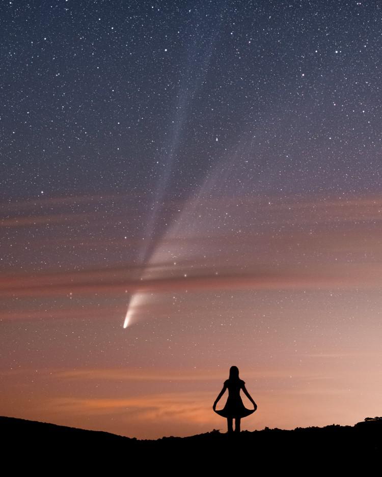 A woman in silhouette appears to greet a comet that appears behind bands of light cloud