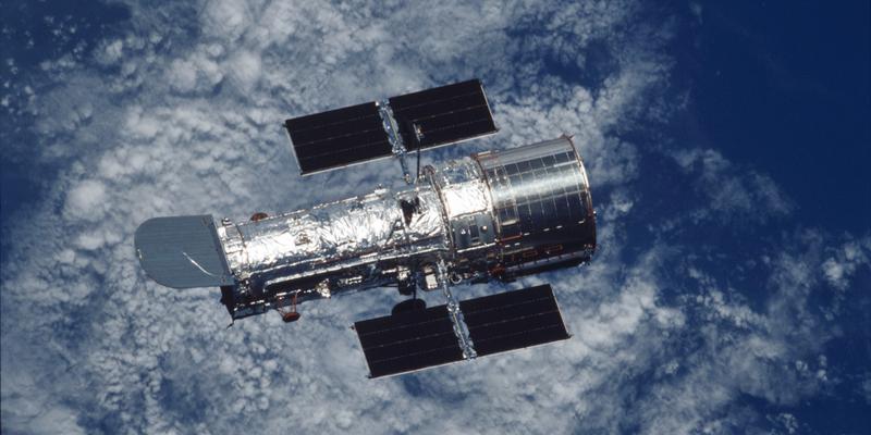 The silver-coloured Hubble Space Telescope with the blue ocean and white clouds of the Earth visible underneath.