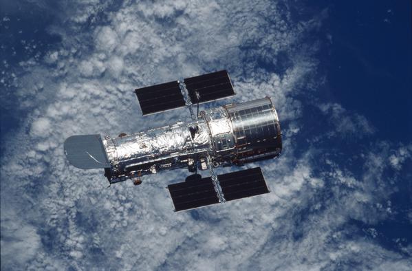 Il telescopio spaziale Hubble, argenteo, con l'oceano blu e le nuvole bianche della Terra visibili al di sotto .