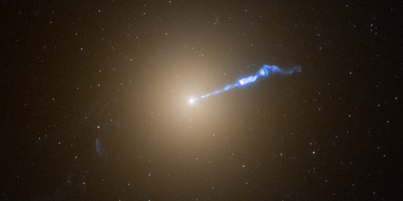 Una galassia rotonda e sfocata, con un nucleo luminoso, ha un getto bianco o blu a macchie che si estende in alto a destra.