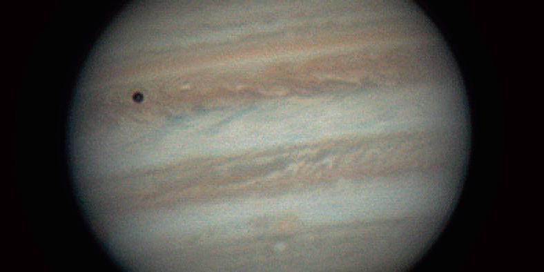 Jupiter with coloured horizontal bands of clouds. The shadow of the moon Io is seen as a dark circle in the top left