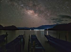 Four boats moored on a lake with the glow of a distant town. The Milky Way with dark and light patches dominates the sky