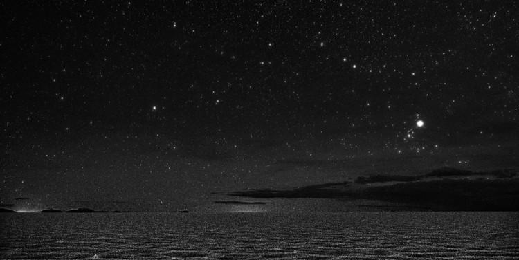 Above a flat, cracked landscape, Orion is shaped like a bow tie. Just above the horizon is a diffuse cluster of bright stars.