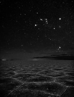 Above a flat, cracked landscape, Orion is shaped like a bow tie. Just above the horizon is a diffuse cluster of bright stars.