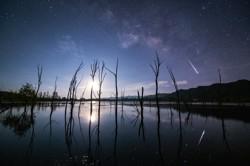 The bright Moon is reflected in a pool of water. The diffuse light and dark patches of the Milky Way dominate the top right