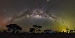 صورة  مجرة درب التبانة  مقوسة فوق أرض عشبية أفريقية. ويظهر توهجها المنتشر متقاطعا مع تيار من البقع الداكنة.