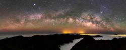 Das diffuse Leuchten der Milchstraße als Bogen mit dunklen Flecken über einer Gebirgskette, die aus den Wolken hervorschaut.