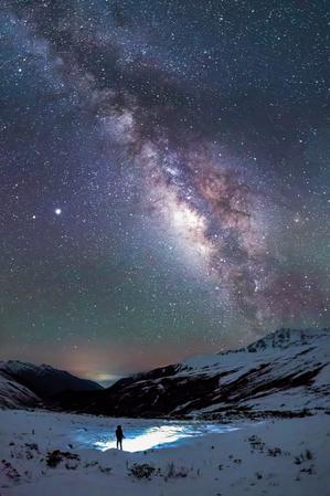 A person stands next to a snowy lake. The Milky Way stretches from the top of the image to the horizon.