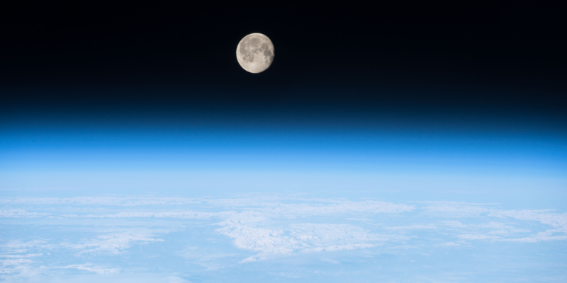 The Moon in the far background and Earth's the blue of the Earth's atmosphere smoothly changes to the black of space