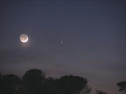 The crescent Moon sits to the left of two bright planets. On the right side the Pleiades star cluster can be seen.