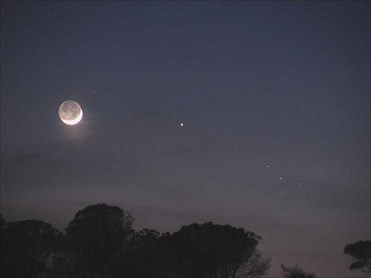The crescent Moon sits to the left of two bright planets. On the right side the Pleiades star cluster can be seen.