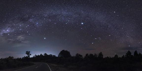 The curve of the Milky Way over a road. Bottom left, two bright stars form a line pointing to a kite-shaped stellar assembly