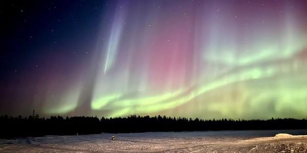 Ripples of green light over a tree-lined horizon. Pillars of white light rise into the sky.