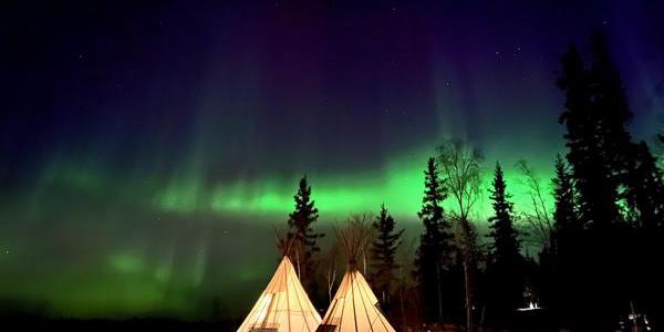 Two illuminated teepees in a subarctic forest. Green bands of aurorae light up the sky.