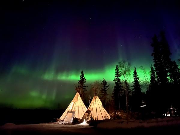 Due teepee illuminati in una foresta subartica. Fasce verdi di aurore illuminano il cielo.