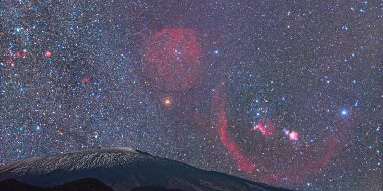 Above a volcano, a bow-tie-shaped Orion is peppered with bright sweeps of nebular gas