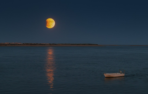 The moon is partially covered by the earth's shadow and shrouded in orange light over beautiful coastal scenery.