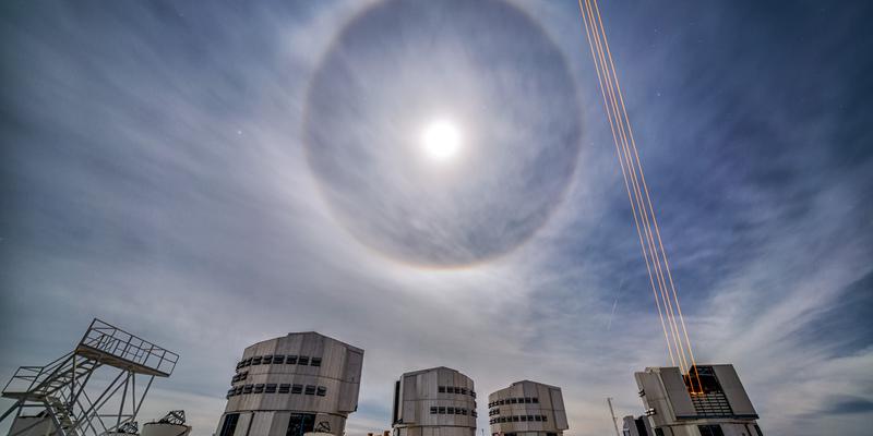 Le quattro cupole del telescopio VLT. Una cupola è aperta e spara raggi laser. La luna in cielo ha un anello di luce intorno.