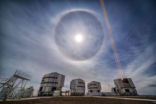 The four telescope domes of VLT. One dome is open and firing laser beams. In the sky, the moon has a ring of light around it.