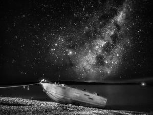 A small boat sits on the shore with the light and dark patches of the Milky Way above