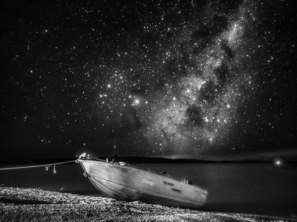 A small boat sits on the shore with the light and dark patches of the Milky Way above