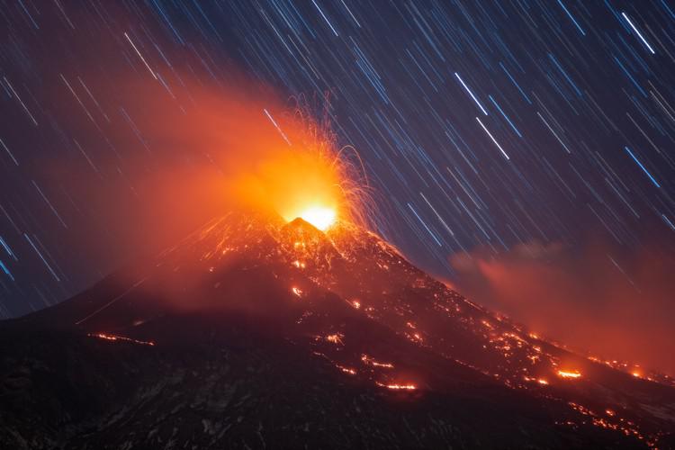 Star trails form short, diagonal lines in the background of a volcanic eruption