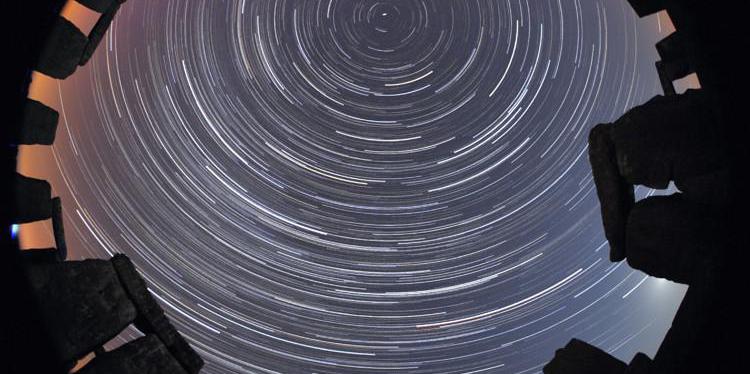 An all-sky image. Above a ring of stones shaped like door-frames, the bright curved paths of star trails seem to form circles