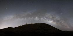 Mehrere Sternwartenkuppeln auf einer Bergkette mit der sich wölbenden Milchstraße im Hintergrund.